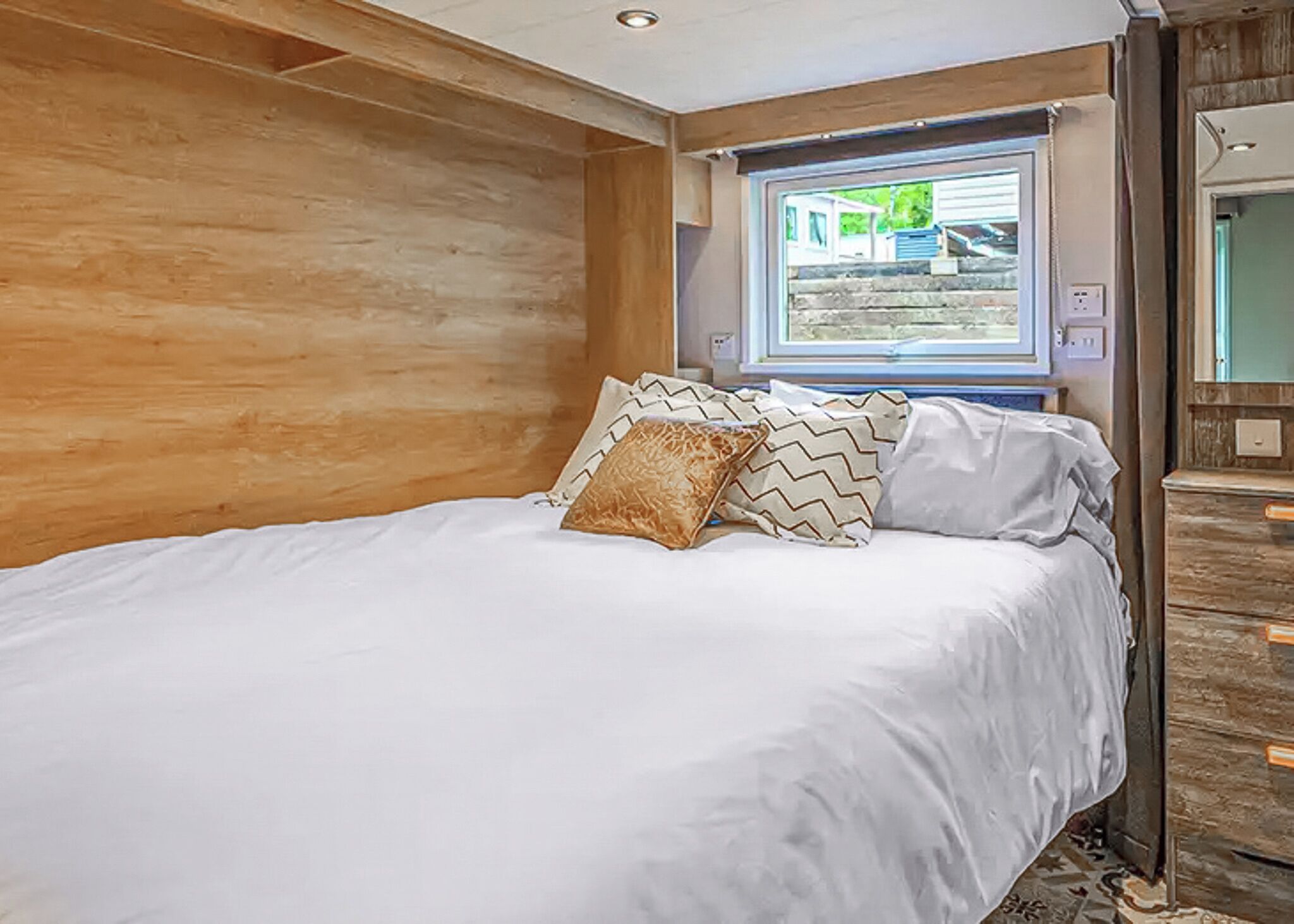 Quarto com parede de madeira, cama com roupa de cama branca, almofadas estampadas, janela com vista e cômoda de madeira.