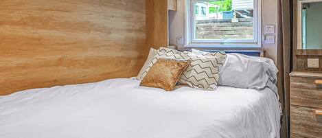 Quarto com parede de madeira, cama com roupa de cama branca, almofadas estampadas, janela com vista e cômoda de madeira.