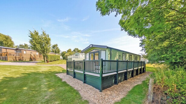 A single-story mobile home with a fenced deck area, surrounded by a well-maintained lawn and trees. - 2 bedroom accommodation in Carnforth (Carnforth)