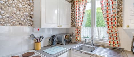 Une cuisine avec un mur en pierre, une horloge, des meubles blancs, une cuisinière, un four à micro-ondes et une fenêtre agrémentée de rideaux à motifs.