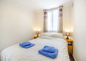 Ein Schlafzimmer mit einem Bett, zwei Nachttischen, einem Fenster mit Vorhängen und zwei gefalteten Handtüchern.
