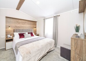 Un dormitorio moderno con cabecera de madera, una cama grande con muchos almohadones, una mesita de noche con una lámpara y una mesita auxiliar con un marco de fotos.