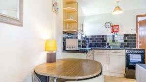 Een kleine keuken met een eettafel, stoelen, een magnetron, een fornuis en een klok aan de muur.