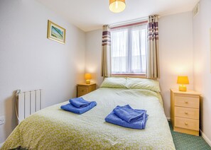 A bedroom with a bed, two bedside tables, a window with curtains, a radiator, and a picture on the wall.
