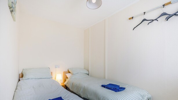 Ein kleines, sauberes Zimmer mit zwei Einzelbetten, einem Nachttisch mit Lampe und einem Kleiderständer mit Kleiderbügeln.