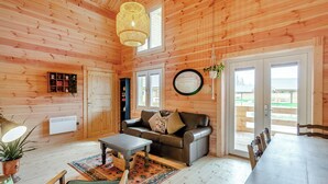 A wooden cabin interior with a sofa, dining table, chairs, a hanging light fixture, a mirror, and a window.