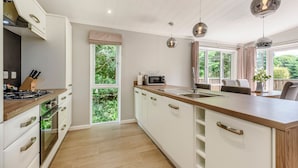 Cocina moderna con armarios blancos, encimera de madera y horno integrado. Hay dos lámparas colgantes, una ventana con vistas a zonas verdes y una zona de comedor con mesa y sillas.