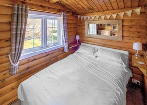 A wooden cabin bedroom with a bed, a window with curtains, a mirror, and a lamp on a bedside table.