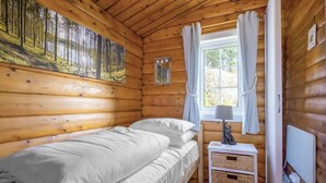 Una habitación de cabaña de madera con una cama, una ventana con cortinas, una mesita de noche con lámpara y un cuadro en la pared.