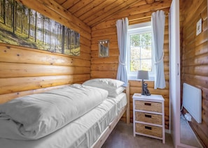 Ein Zimmer in einer Holzhütte mit einem Bett, einem Fenster mit Vorhängen, einem Nachttisch mit Lampe und einem Gemälde an der Wand.