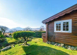 A wooden cabin with a stone foundation, a small porch, and a well-maintained garden with a hedge and a tree.