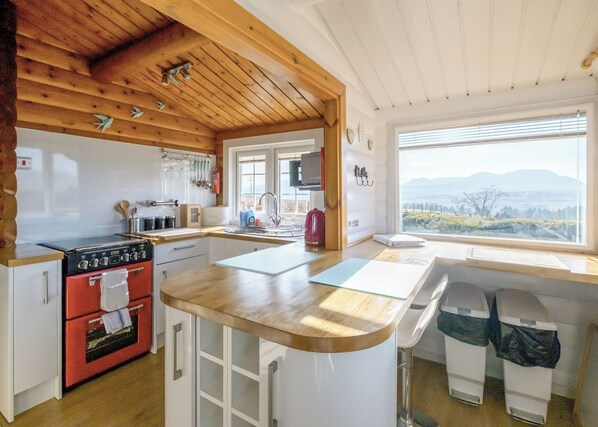 A kitchen with a wooden ceiling, a red stove, a sink, and a window with a view of the countryside.