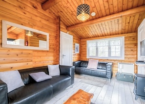 A log cabin interior with a black leather sofa, a wooden coffee table, a fireplace, and a large window.