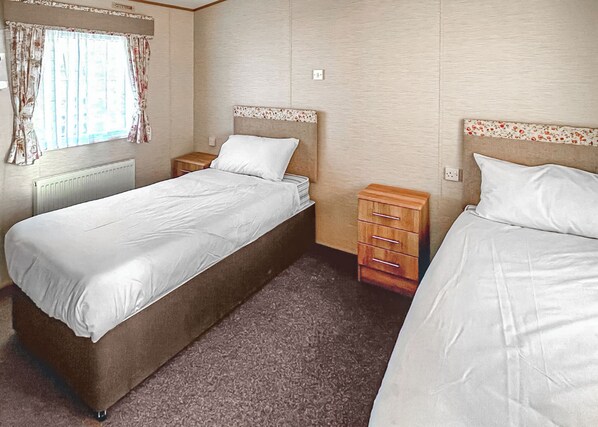A bedroom with two beds, a window with curtains, a radiator, and a wooden nightstand.