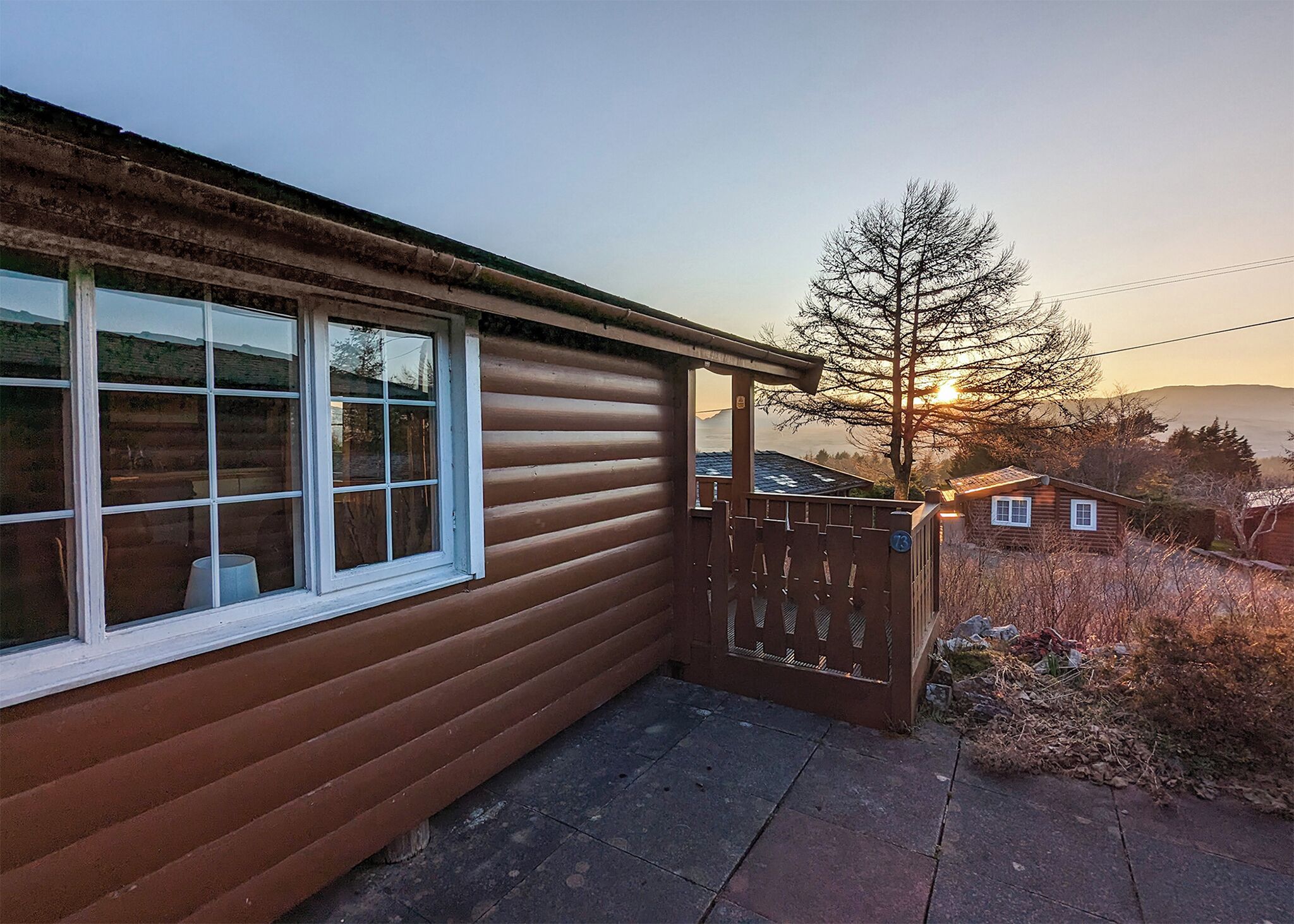 A wooden cabin with a window, a wooden gate, and a view of a tree and mountains in the background.