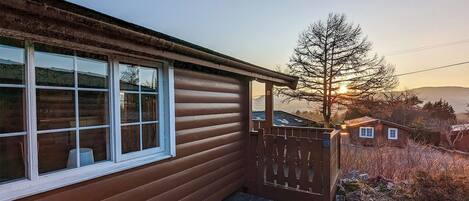 A wooden cabin with a window, a wooden gate, and a view of a tree and mountains in the background.