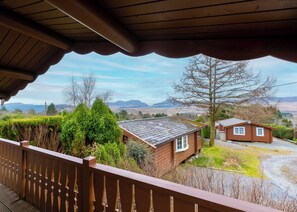 A wooden cabin with a sloped roof, surrounded by greenery and a view of distant mountains.