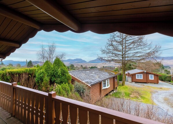 A wooden cabin with a sloped roof, surrounded by greenery and a view of distant mountains.