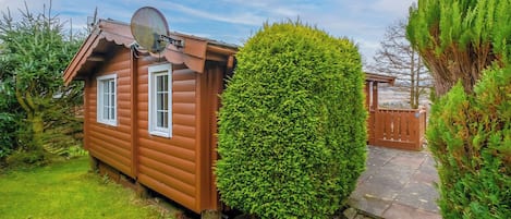 Une petite cabane en bois, dotée d’une antenne parabolique, entourée d’une végétation luxuriante et bordée d’un sentier en pierre.