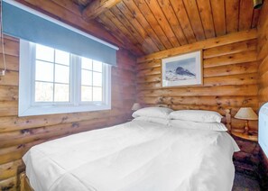 Ein Schlafzimmer in einer Blockhütte mit einem Bett, einem Fenster, einem Bild an der Wand und einer Lampe.