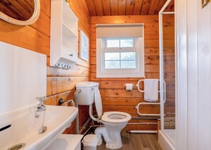 Pieni, siisti kylpyhuone, joka on rakennettu puusta. Siellä on valkoinen wc-istuin, pesuallas hanalla, peili ja ikkuna.
