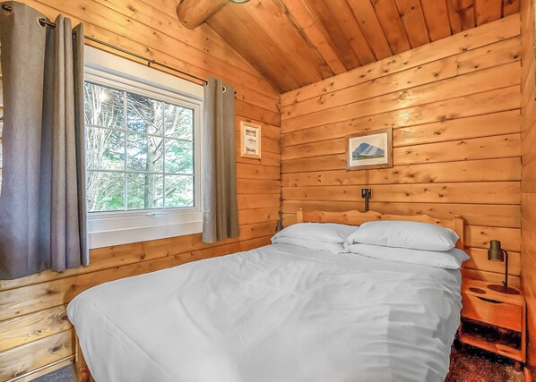 A wooden cabin bedroom with a bed, window, curtains, and a framed picture on the wall.