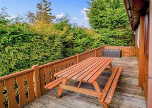 Une terrasse en bois avec une table de pique-nique, entourée d’une végétation luxuriante et d’arbres.