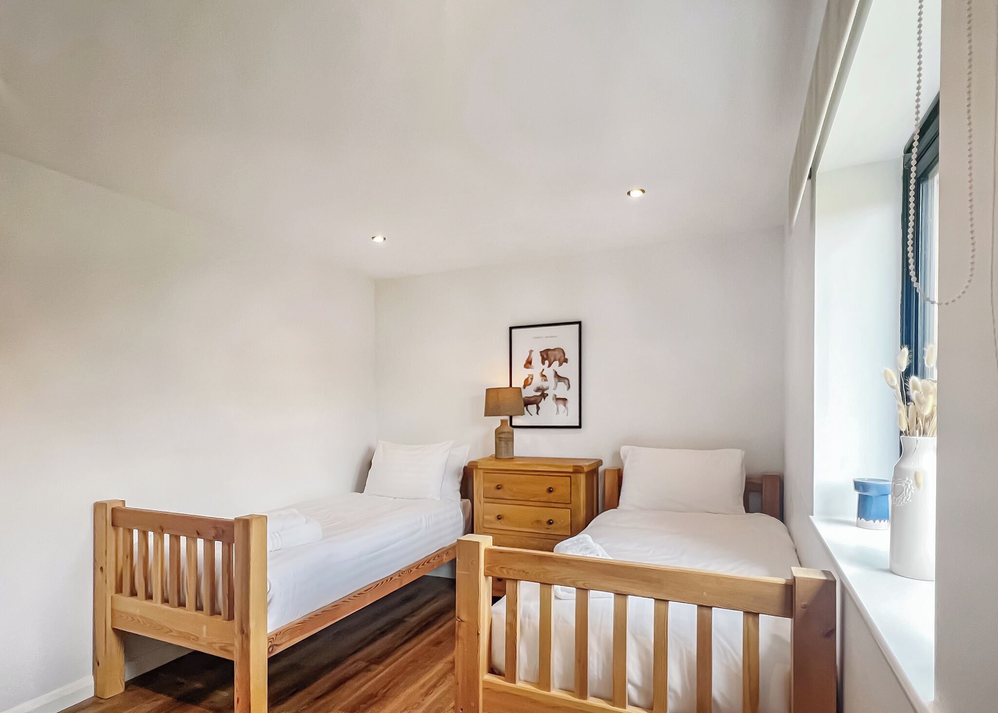 A bedroom with two wooden beds, a nightstand with a lamp, a framed animal illustration, and a window with a view.