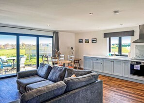 A modern living room with a grey sofa, dining area, kitchen, and large windows offering a view of the outdoors. - 2 bedroom accommodation in Old Buckenham (Old Buckenham)