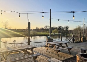 A wooden deck with picnic tables and string lights, overlooking a calm body of water with a distant view of houses and trees. - 2 bedroom accommodation in Old Buckenham (Old Buckenham)