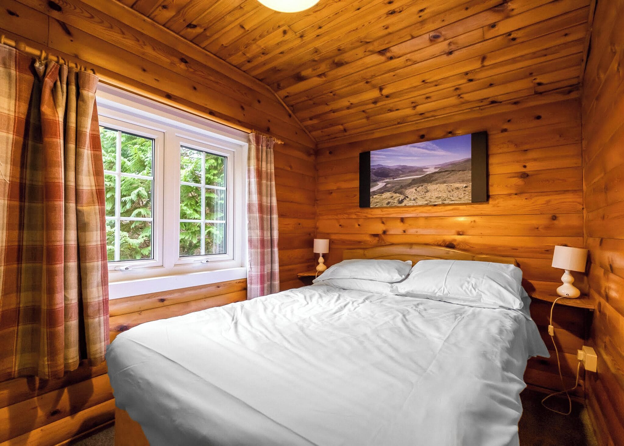 A wooden cabin room with a bed, two bedside lamps, a window with curtains, and a landscape painting on the wall.