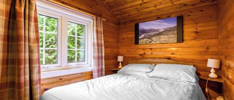 Ein Zimmer in einer Holzhütte mit einem Bett, zwei Nachttischlampen, einem mit Vorhängen versehenen Fenster und einem Landschaftsbild an der Wand.