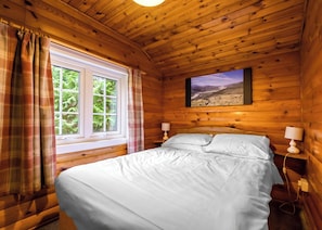 Ein Zimmer in einer Holzhütte mit einem Bett, zwei Nachttischlampen, einem mit Vorhängen versehenen Fenster und einem Landschaftsbild an der Wand.