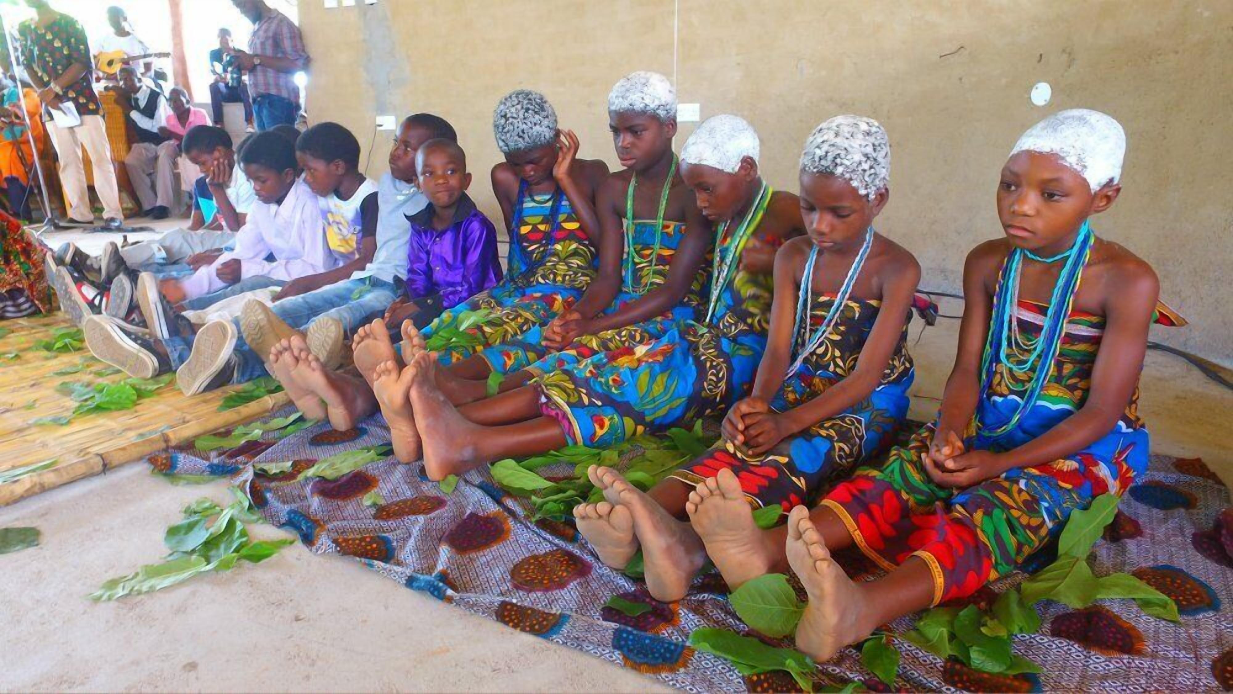 A group of children sitting on a patterned mat in a simple room with a concrete floor and plain walls.