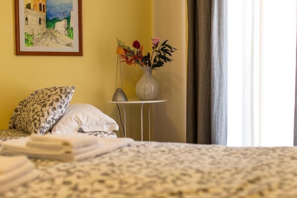 Un letto rifatto con cura, lenzuola a fantasia, un comodino con una lampada, un vaso con fiori e un quadro appeso al muro.