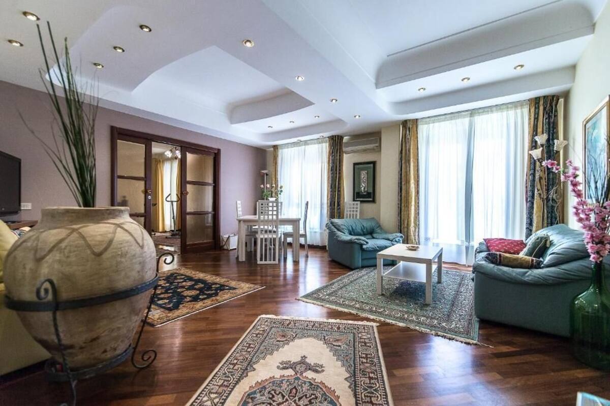 Uma sala de estar ampla com sofá, mesa de centro e um vaso grande. Há uma área de jantar com mesa e cadeiras, e uma janela grande com cortinas.