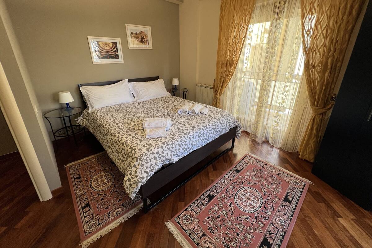 Um quarto com uma cama, duas mesas de cabeceira, uma janela com cortinas e duas imagens penduradas na parede.
