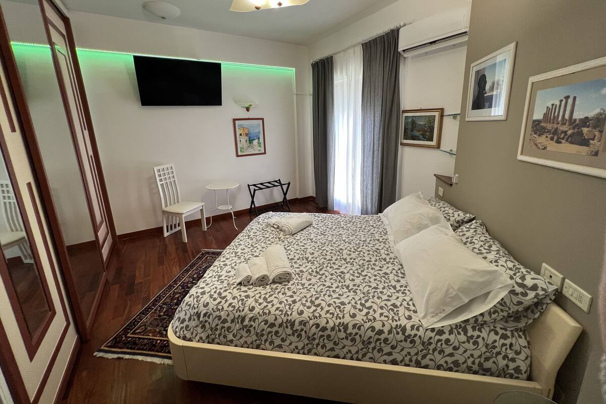 Quarto com cama, escrivaninha, cadeira, televisão e janela com cortinas.