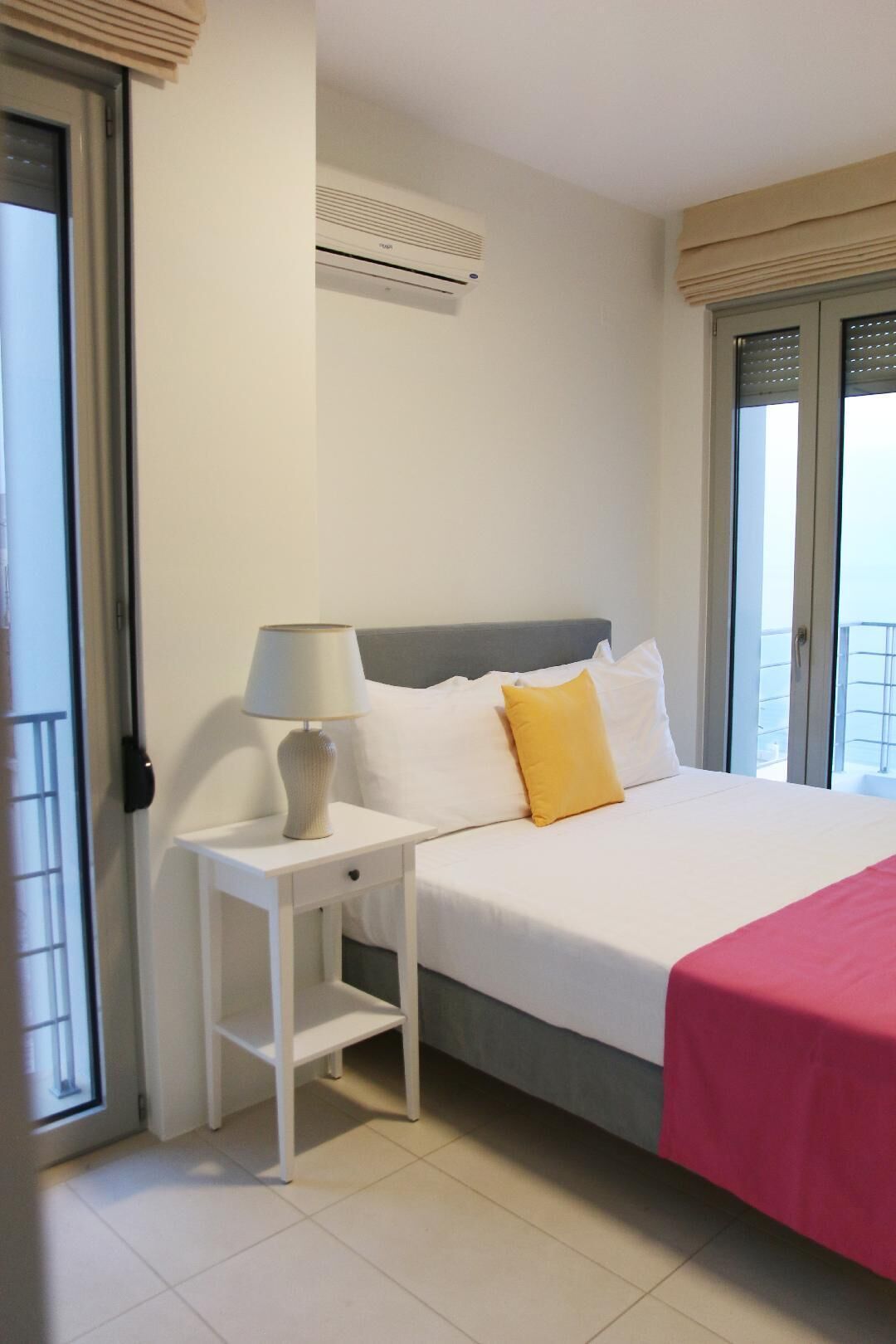 Una habitación de hotel con una cama, una mesita de noche con lámpara, una ventana con vistas y un equipo de aire acondicionado.