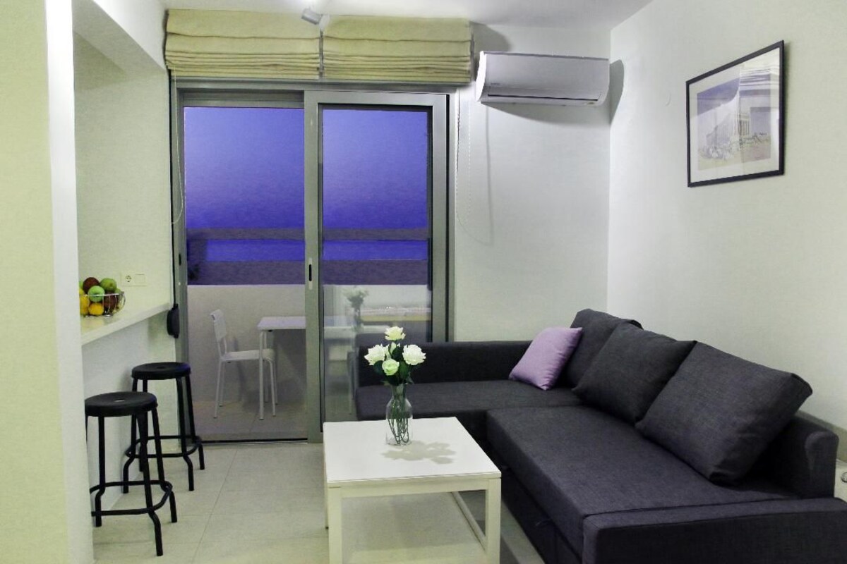 A modern living room with a dark grey sofa, a white coffee table, and a vase with white flowers. There is a glass door leading to a balcony with a view of the sky.