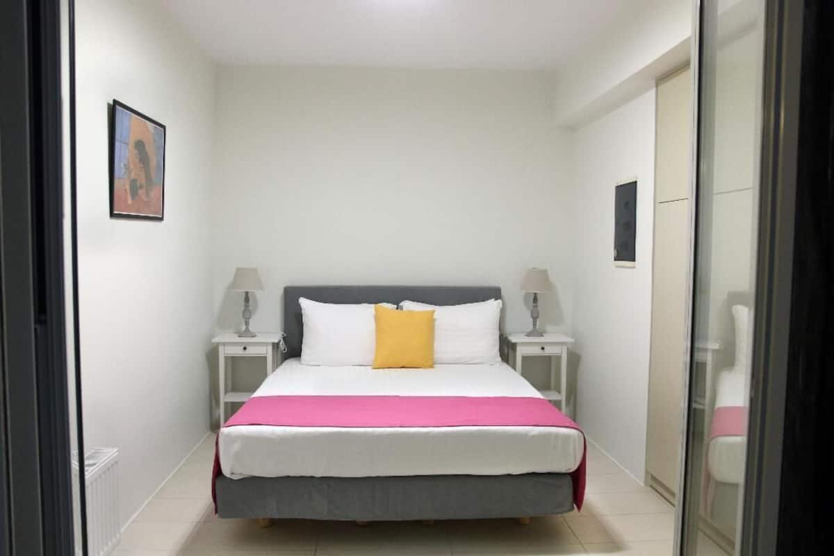 Une chambre d’hôtel étroite, avec un lit, deux tables de chevet, une petite table, une couvre-lit rose et un seul oreiller jaune.