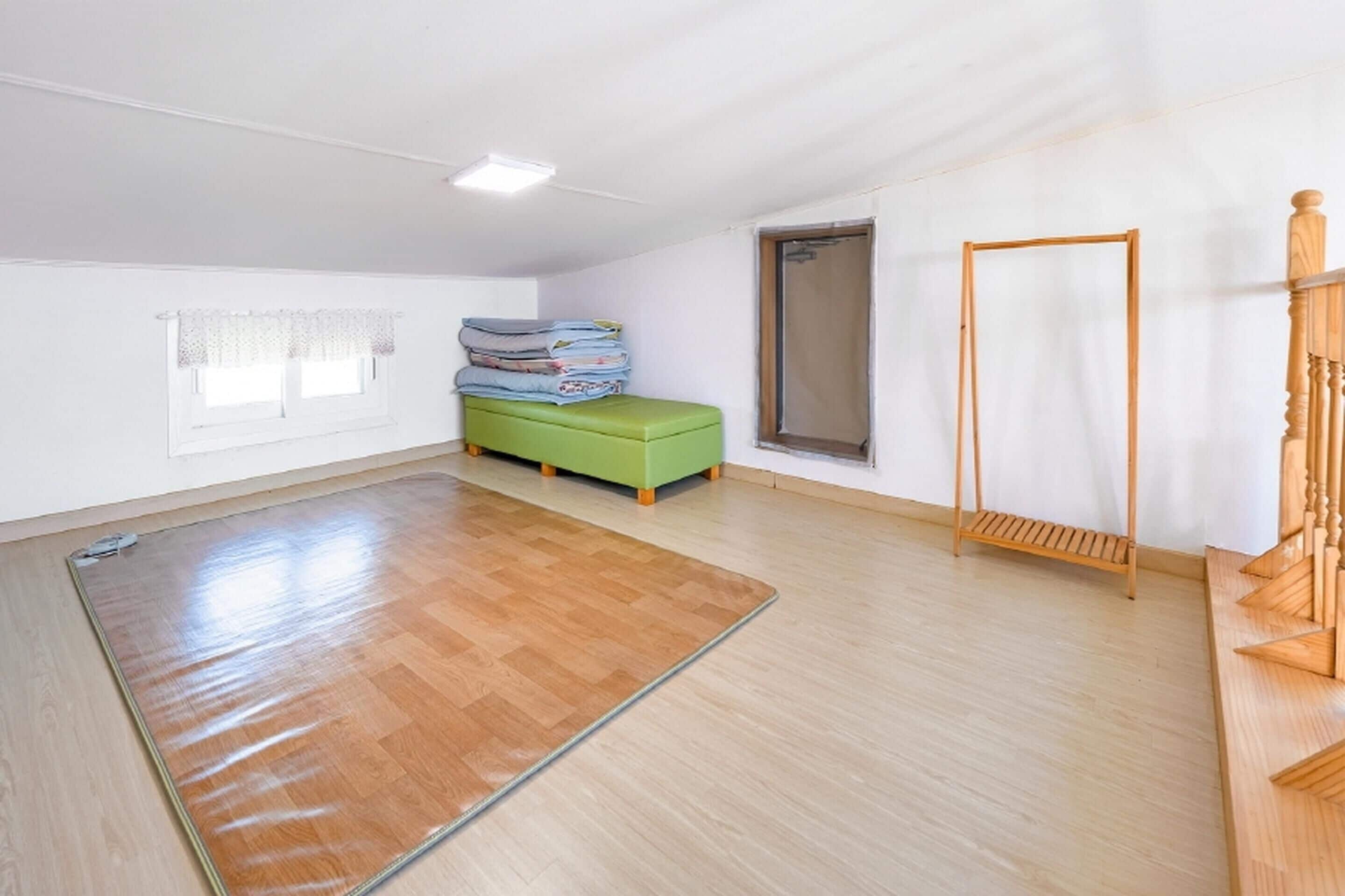 Ein geräumiger Raum mit Holzboden, einer grünen Bank, einer Holzleiter und einem Fenster mit weißem Vorhang.