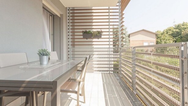 Un balcon avec une table et des chaises en verre, une rambarde en métal et un pot de fleurs.