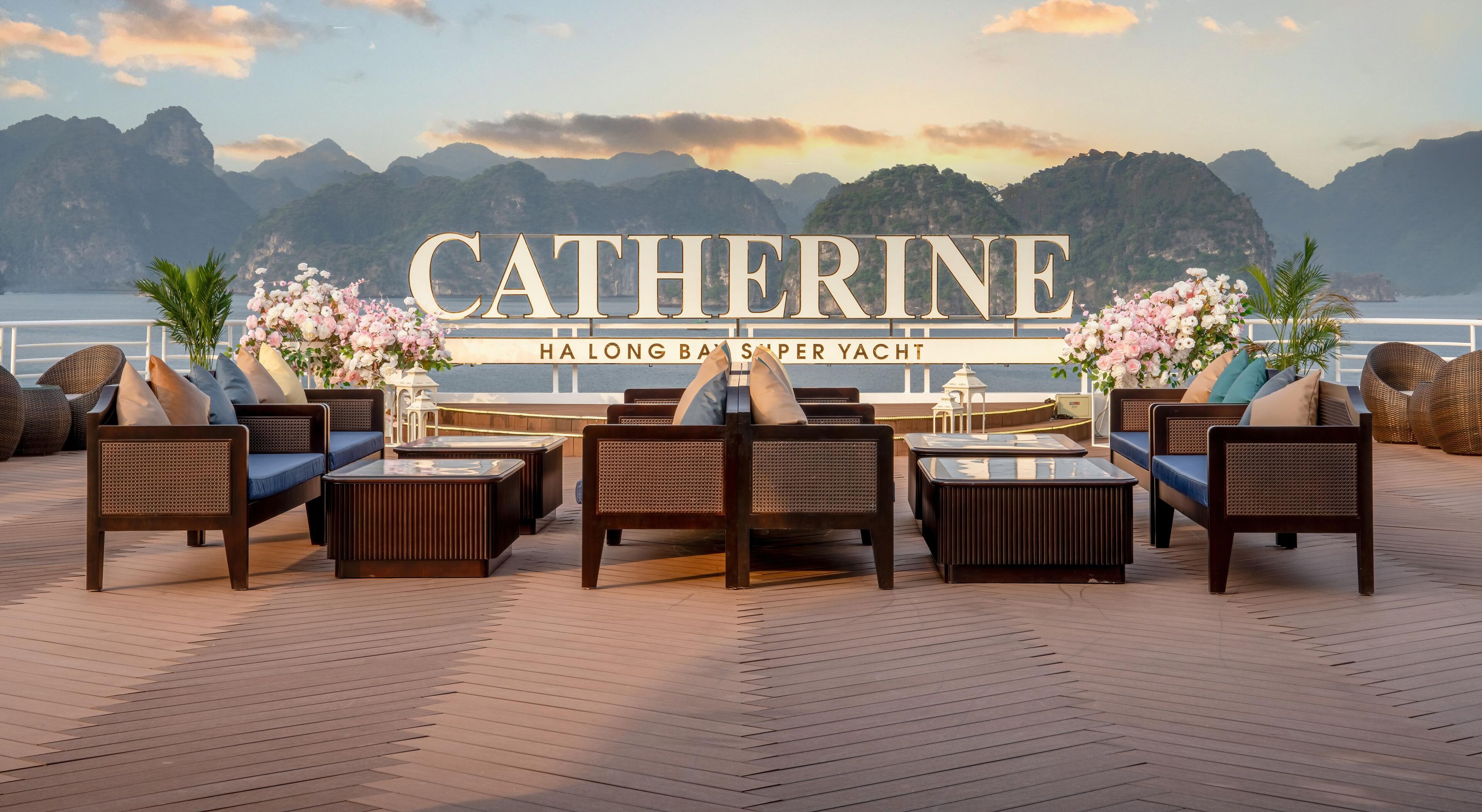 A yacht deck with a lounge area featuring sofas and tables, a large sign reading "CATHERINE HA LONG BAY SUPER YACHT", and a backdrop of mountains and water.