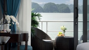 Une pièce dotée d’une grande porte-fenêtre coulissante donnant sur un balcon avec vue sur la mer et les montagnes. On y trouve un lit, une petite table avec un vase de fleurs et des meubles en osier.