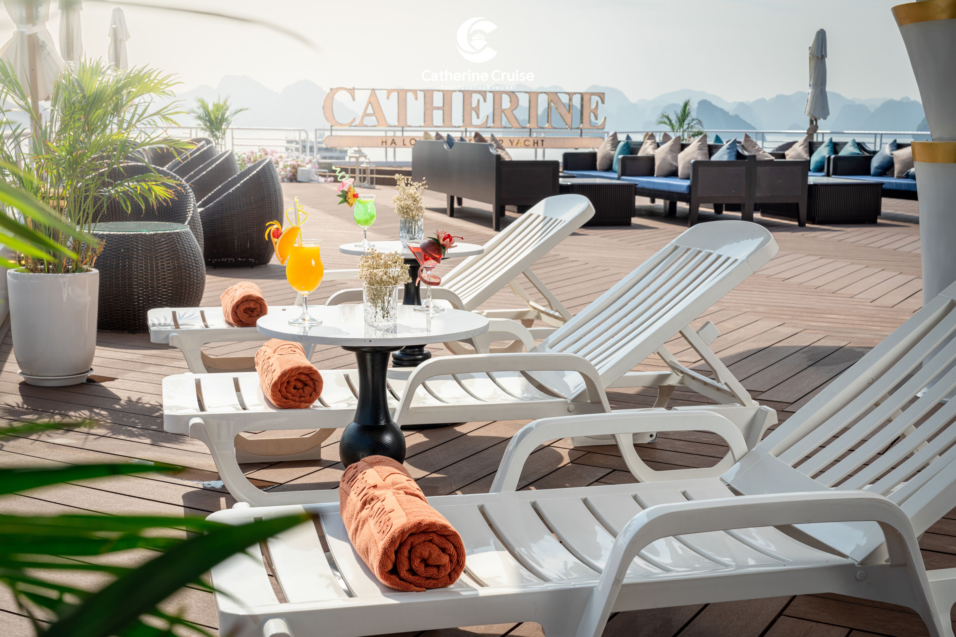 A sunlit outdoor lounge area with white lounge chairs, a table with drinks and towels, and a view of the sea and mountains in the background.