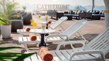 A sunlit outdoor lounge area with white lounge chairs, a table with drinks and towels, and a view of the sea and mountains in the background.
