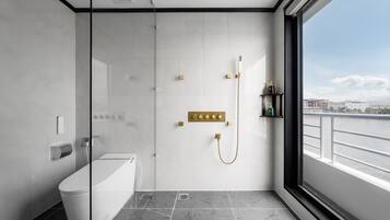 A modern bathroom with a bathtub, a shower with gold fixtures, and a view of a river through a large window.