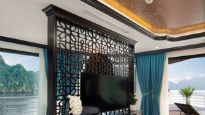 A room with a TV on a wooden cabinet, a large window with a view of a body of water, and a balcony with a chair and plant. - Catherine Cruises (Tuan Chau)