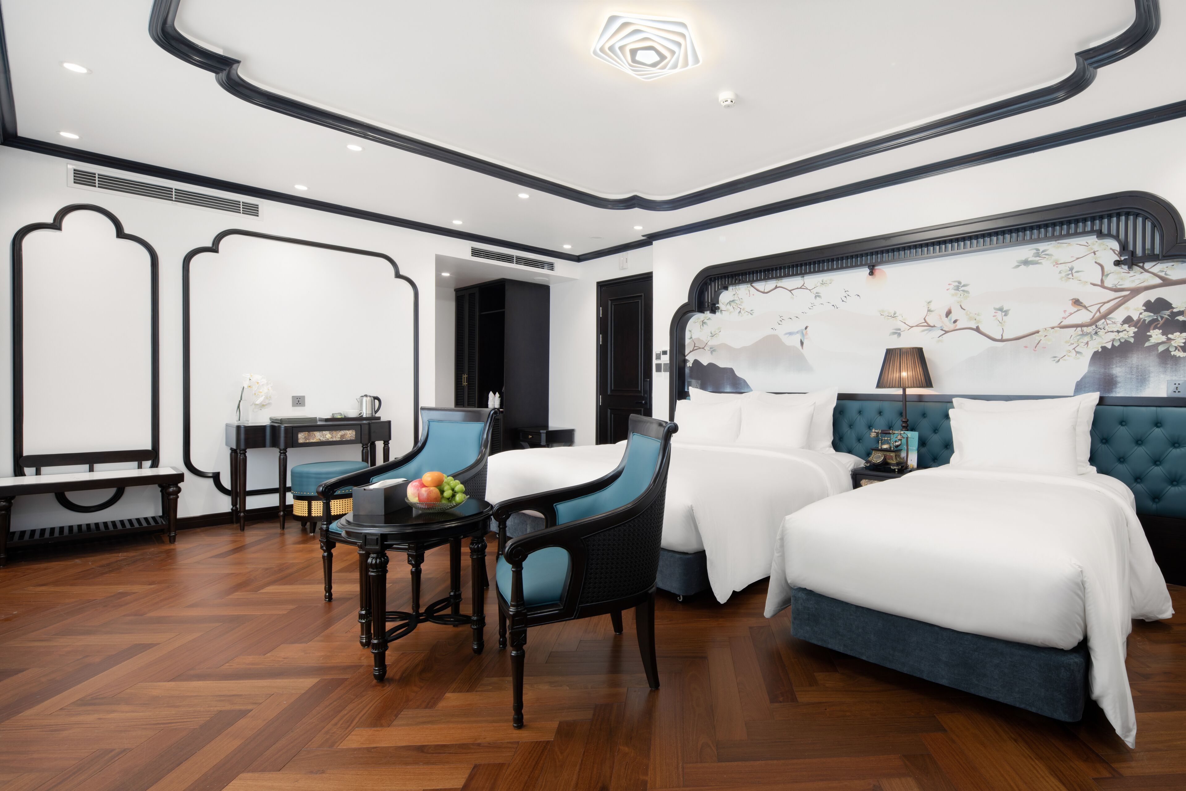 A hotel room with a large bed, two chairs, a small table with fruit, and a decorative wall mural.